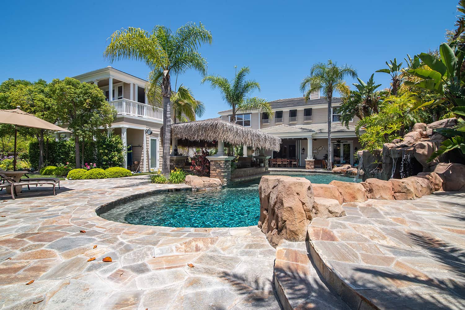 Luxury home backyard with palapa top swim up bar, pool, rock slide, and palm trees.